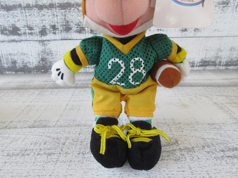 May include: A plush toy of a character dressed in a green and yellow football uniform, holding a brown football. The jersey has the number 28 in white. The character wears black shoes with yellow laces.