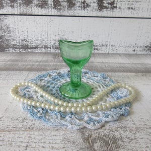 Puede incluir: Una copa ocular de vidrio verde claro translúcido con un borde curvo y una base de pedestal resistente, acompañada por un collar de cuentas de perlas blancas artificiales sobre un tapete de encaje de ganchillo azul y blanco.