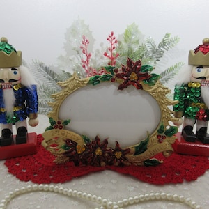 Puede incluir: Un marco de fotos de color dorado con una abertura ovalada blanca, decorado con poinsettias rojas, acebo verde y las palabras "Feliz Navidad". Dos figuras de cascanueces, una en lentejuelas azules y verdes y otra en lentejuelas verdes y doradas, se colocan a cada lado del marco.