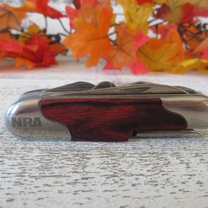 May include: A multi-tool knife with a stainless steel and wood grain design. The knife has the letters "NRA" on the side. The background features fall leaves.