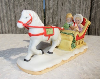 May include: A white porcelain horse pulling a yellow and red sleigh with two people inside. The people are wearing winter clothing and are holding a basket. The sleigh is decorated with green holly.