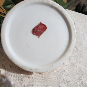 May include: The bottom of a white ceramic figurine with a red and gold label that reads "Lefton China".