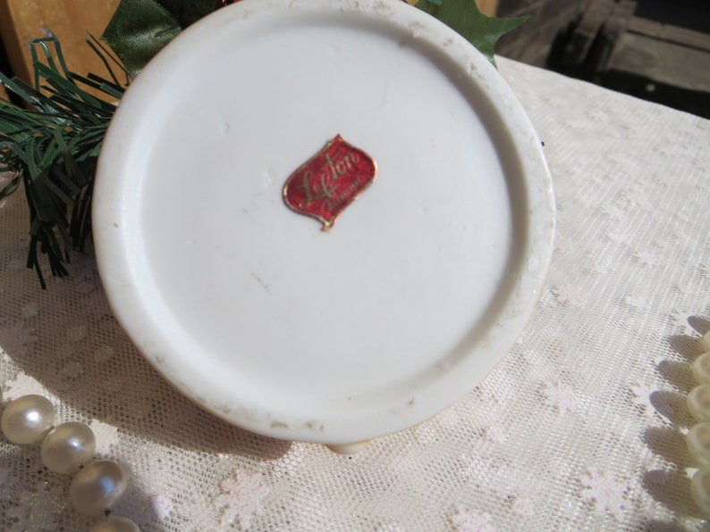 May include: The bottom of a white ceramic figurine with a red and gold label that reads "Lefton China".