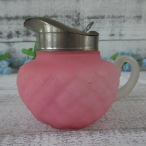May include: A small pink frosted glass creamer with a diamond-patterned texture. The pitcher features a silver-toned metal lid with a hinged spout and a curved, translucent glass handle attached to the side of the rounded, bulbous body.