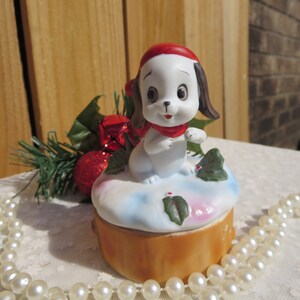 May include: A white porcelain figurine of a dog wearing a red Santa hat and scarf, sitting on a snow-covered log with holly leaves. The figurine is part of a musical trinket box.