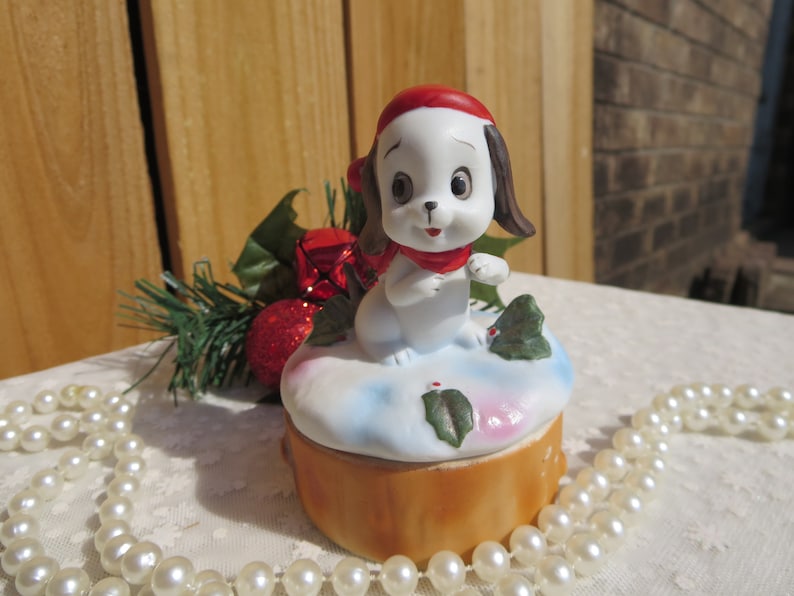 May include: A white porcelain figurine of a dog wearing a red Santa hat and scarf, sitting on a snow-covered log with holly leaves. The figurine is part of a musical trinket box.