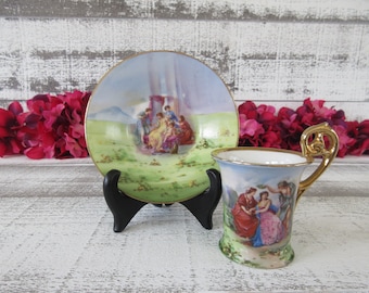 May include: A vintage porcelain teacup and saucer set with a gold trim. The saucer features a painted scene of people in a garden setting. The teacup has a similar scene of people in a garden setting.