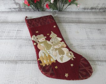 May include: A burgundy Christmas stocking with a snowman design. The stocking features a textured fabric with gold and white appliqués of snowmen, a fence, and stars. The top has a patterned design.