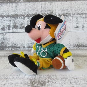May include: A plush Mickey Mouse toy wearing a green and yellow football uniform and helmet. The toy is holding a brown football. The jersey has the number 16 on it. The toy has black ears, a pink face, and a red mouth.
