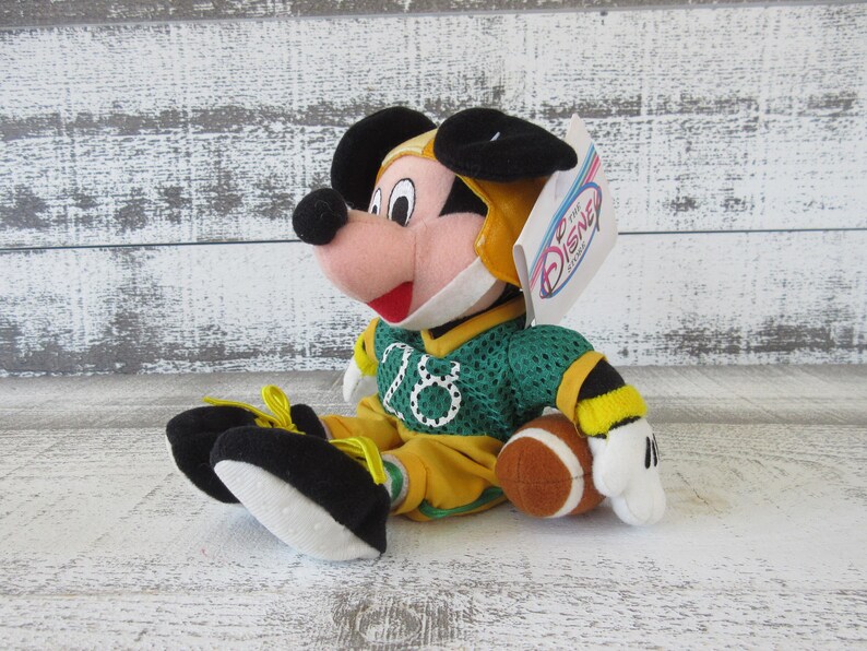 May include: A plush Mickey Mouse toy wearing a green and yellow football uniform and helmet. The toy is holding a brown football. The jersey has the number 16 on it. The toy has black ears, a pink face, and a red mouth.