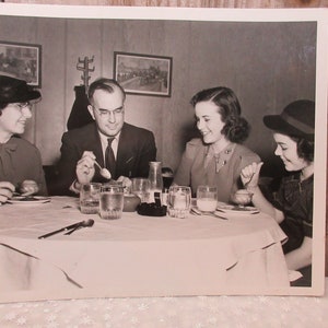 Puede incluir: Una fotografía en blanco y negro de cuatro personas sentadas a una mesa. Las personas sonríen y parecen disfrutar de una comida juntos. Hay vasos de agua y leche en la mesa, así como algunos platos de comida.