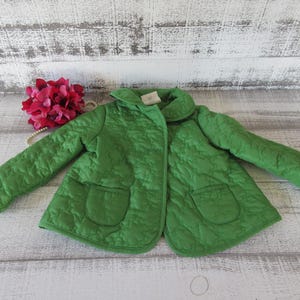 Peut inclure: Une veste matelassée verte avec deux poches avant et un col arrondi. La veste est d'une nuance de vert vif et semble être faite d'un tissu doux et texturé. La veste est présentée sur une surface en bois de couleur claire.