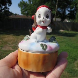 May include: A white porcelain trinket box with a small, white dog figurine wearing a red hat and scarf. The box is decorated with a blue and pink floral design and has a brown wooden base.