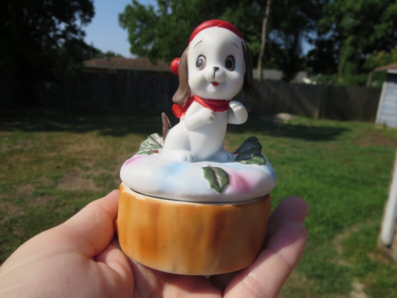 May include: A white porcelain trinket box with a small, white dog figurine wearing a red hat and scarf. The box is decorated with a blue and pink floral design and has a brown wooden base.