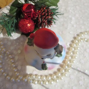May include: A white ceramic Christmas bell with a red Santa hat and a brown dog's head peeking out from under the hat. The bell is decorated with holly and snowflakes. The bell is surrounded by a string of white pearls.