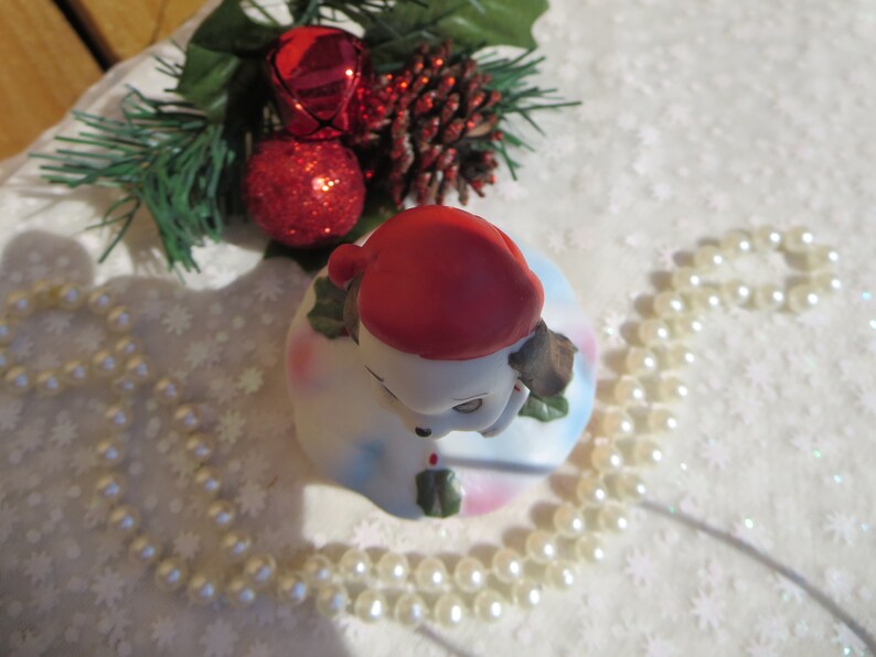 May include: A white ceramic Christmas bell with a red Santa hat and a brown dog's head peeking out from under the hat. The bell is decorated with holly and snowflakes. The bell is surrounded by a string of white pearls.