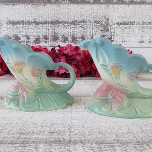 Könnte beinhalten: Zwei Keramikvasen im floralen Design. Die Vasen sind in einem hellblauen, rosa- und grünen Farbschema gehalten. Jede Vase hat einen Griff und ist mit Blumen verziert. Die Vasen stehen auf einer weißen Holzoberfläche.