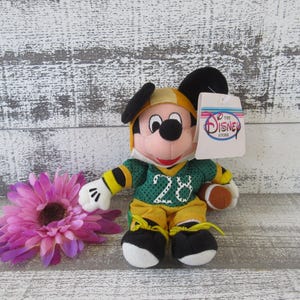 May include: A Mickey Mouse plush toy dressed in a green and yellow football uniform with the number 28. The toy has a football and a tag that says "The Disney Store". A purple flower is in the foreground.