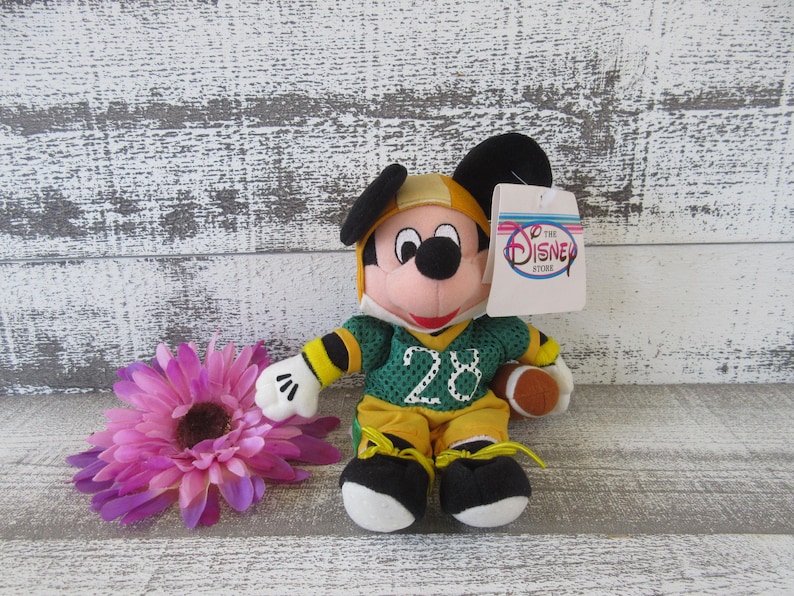 May include: A Mickey Mouse plush toy dressed in a green and yellow football uniform with the number 28. The toy has a football and a tag that says "The Disney Store". A purple flower is in the foreground.