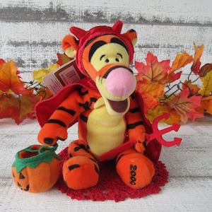 Könnte beinhalten: Ein Plüschtier von Tigger, der sich für Halloween als Teufel verkleidet hat. Der orange und schwarz gestreifte Tiger trägt ein rotes Teufelskostüm, einen Umhang und hält einen roten Dreizack. Ein kleiner orangefarbener Kürbis-Eimer befindet sich in der Nähe.
