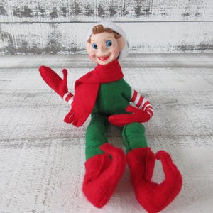 May include: A whimsical elf doll with a smiling face, wearing a green outfit, red scarf, and matching red gloves and shoes. The gloves have white and red striped sleeves. The elf has a white hat and is posed with its arms outstretched.