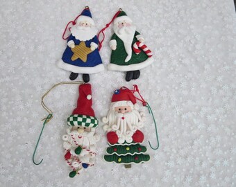 May include: Four handmade Christmas ornaments featuring Santa Claus in various poses. The ornaments are made of a white clay-like material and are painted in bright colors. One Santa is wearing a blue and white suit and holding a gold star. Another Santa is wearing a green suit and holding a candy cane. The third Santa is wearing a red and white suit and has a white beard. The fourth Santa is wearing a red and white suit and has a white beard and a green tree.