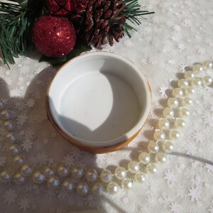 May include: A white ceramic trinket box with a gold rim. The box is empty and has a smooth, white interior. The box is surrounded by a string of white pearls and a pine branch with red ornaments.