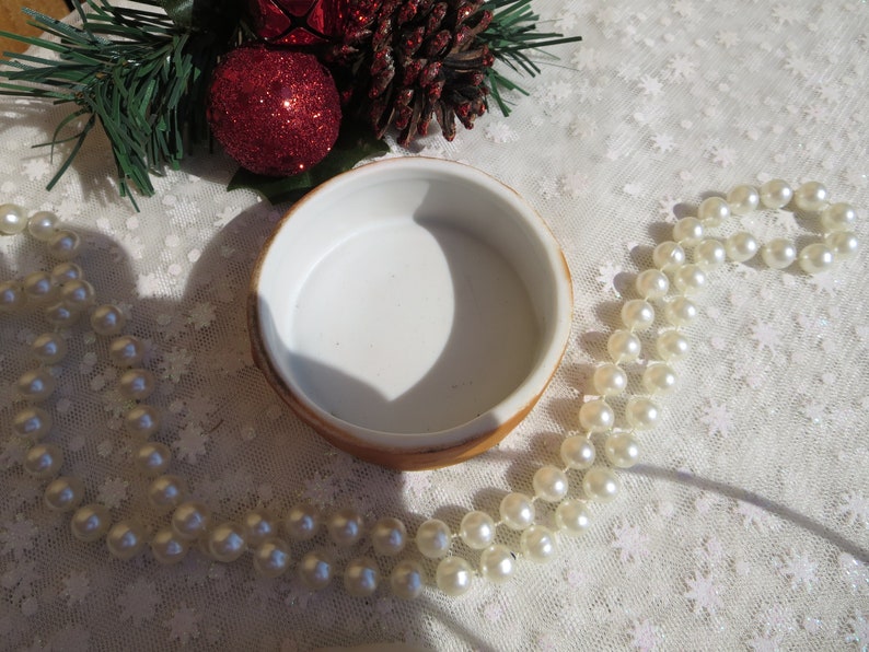May include: A white ceramic trinket box with a gold rim. The box is empty and has a smooth, white interior. The box is surrounded by a string of white pearls and a pine branch with red ornaments.