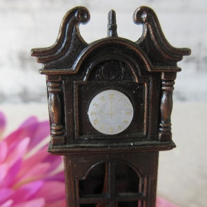 Puede incluir: Un sacapuntas de metal marr&oacute;n de estilo vintage con forma de reloj de pie. La esfera del reloj tiene n&uacute;meros romanos y un fondo blanco.