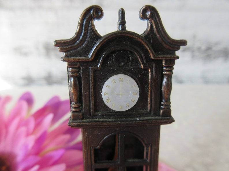 Puede incluir: Un sacapuntas de metal marr&oacute;n de estilo vintage con forma de reloj de pie. La esfera del reloj tiene n&uacute;meros romanos y un fondo blanco.