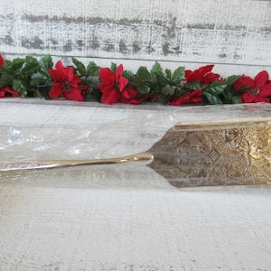 Puede incluir: Una paleta de pastel dorada con un diseño floral grabado y ornamentado. La paleta está en una funda de plástico transparente. Flores de pascua rojas y hojas verdes en el fondo.