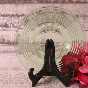 May include: A clear, light green glass plate featuring a series of concentric circular ridges and a textured, grid-like pattern around the outer rim. The plate is displayed upright on a black plastic stand, showcasing its translucent quality.