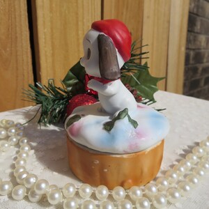 May include: A white ceramic Christmas trinket box with a small dog wearing a red hat and scarf. The box is decorated with a snowy scene and green holly.