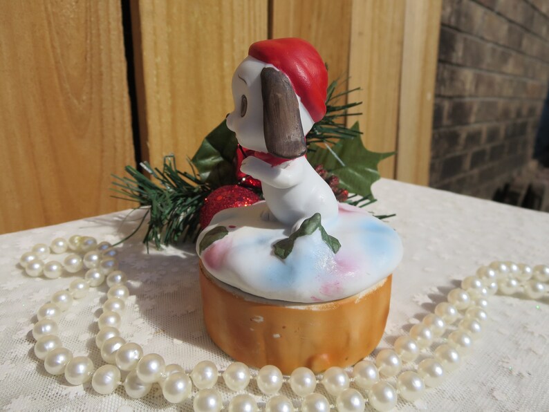 May include: A white ceramic Christmas trinket box with a small dog wearing a red hat and scarf. The box is decorated with a snowy scene and green holly.
