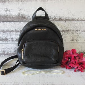 Puede incluir: Una mini mochila de cuero negro granulado con asa superior, correas ajustables para los hombros y un bolsillo frontal con cremallera. El nombre de la marca MICHAEL KORS está impreso en letras doradas en la parte superior frontal del bolso.