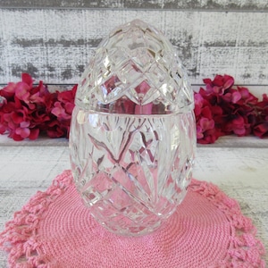 May include: A clear, cut-glass jar with a lid, on a pink crocheted doily. The jar features a diamond pattern on the lid and body. Red flowers are in the background against a white-washed wooden surface.