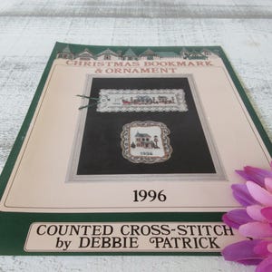 May include: A counted cross-stitch pattern booklet for a Christmas bookmark and ornament. The pattern features a house with a Christmas tree and a sleigh with reindeer. The booklet is titled "Christmas Bookmark & Ornament" and "Counted Cross-Stitch by Debbie Patrick".