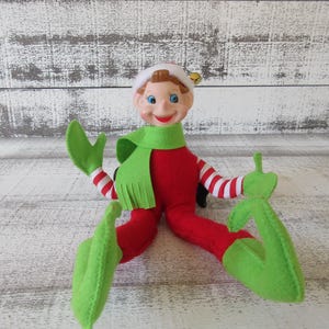 May include: A vintage Christmas elf doll with a smiling face, wearing a red suit, green scarf, and green elf shoes. The elf has white and red striped arms and a white hat with a gold bell.