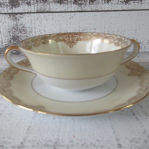 May include: A white porcelain soup bowl with gold trim and a matching saucer. The bowl has two handles and a scalloped edge.