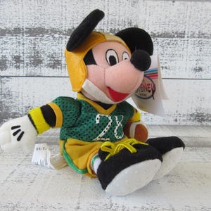 May include: A plush Mickey Mouse toy wearing a yellow football helmet and a green and yellow jersey with the number 28. The toy has black ears, a red mouth, and white gloves. A Disney Store tag is attached.