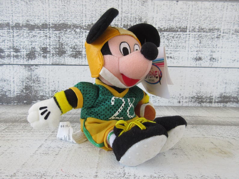 May include: A plush Mickey Mouse toy wearing a yellow football helmet and a green and yellow jersey with the number 28. The toy has black ears, a red mouth, and white gloves. A Disney Store tag is attached.