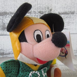 May include: A plush Mickey Mouse toy wearing a yellow and black football helmet and a green jersey with yellow accents. The toy has a pink face, black ears, and a black nose. A tag is attached to the toy.