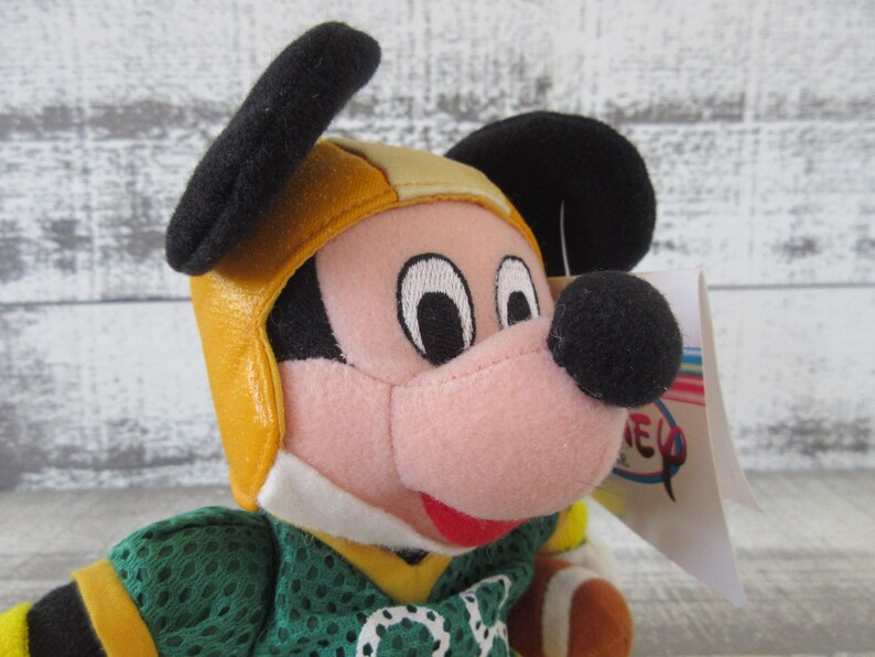 May include: A plush Mickey Mouse toy wearing a yellow and black football helmet and a green jersey with yellow accents. The toy has a pink face, black ears, and a black nose. A tag is attached to the toy.