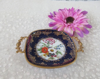 May include: A small, square, blue and gold tray with a floral design and gold handles. The tray is on a white lace tablecloth.