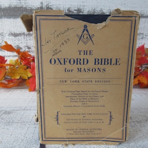 May include: The Oxford Bible for Masons, New York State Edition, in a worn cardboard box. The cover is tan with blue text and a Masonic symbol. Handwritten inscription reads "Jay Taylor Jan 1957".