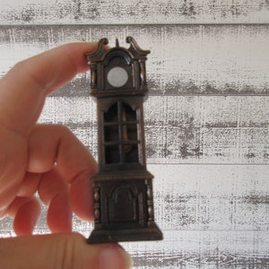 Puede incluir: Un peque&ntilde;o sacapuntas de metal de estilo antiguo con forma de reloj de pie. El sacapuntas es marr&oacute;n y tiene una esfera blanca con una aguja negra.