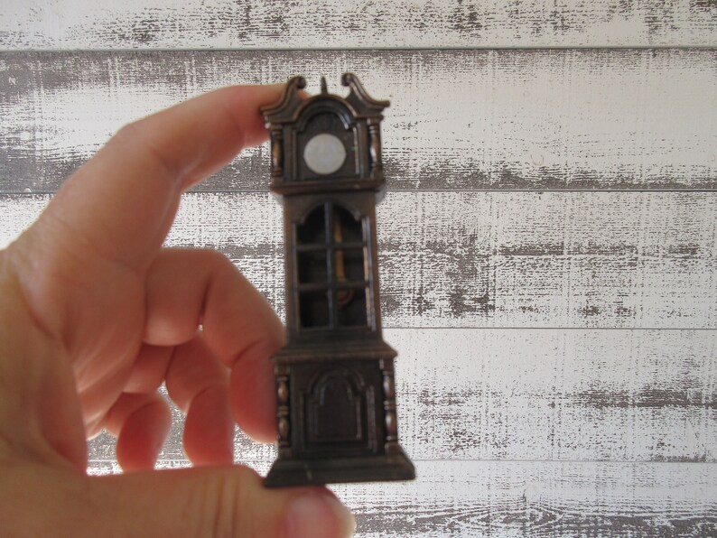 Puede incluir: Un peque&ntilde;o sacapuntas de metal de estilo antiguo con forma de reloj de pie. El sacapuntas es marr&oacute;n y tiene una esfera blanca con una aguja negra.