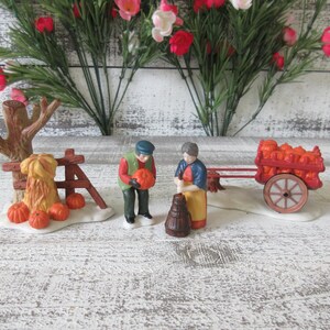 Puede incluir: Una escena otoñal en miniatura con figuras de granjeros, calabazas y un carro. La escena incluye una valla de madera, un árbol y un carro lleno de calabazas. Las figuras visten atuendos otoñales y las calabazas son naranjas.