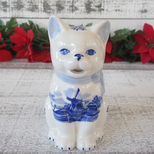 May include: A white ceramic cat figurine with blue accents. The cat has blue eyes, a blue collar, and a blue and white windmill design on its chest. The figurine is sitting upright.