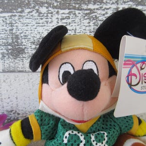 May include: A Mickey Mouse plush toy wearing a green football jersey with the number 00. The toy has a yellow helmet and black ears. A tag from The Disney Store is attached.